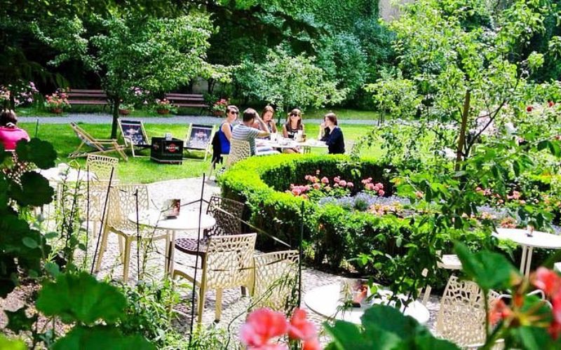 Zdjęcie przedstawia ogródek Meho Cafe Bar Garden, w którym siedzą ludzie w otoczeniu roślinności