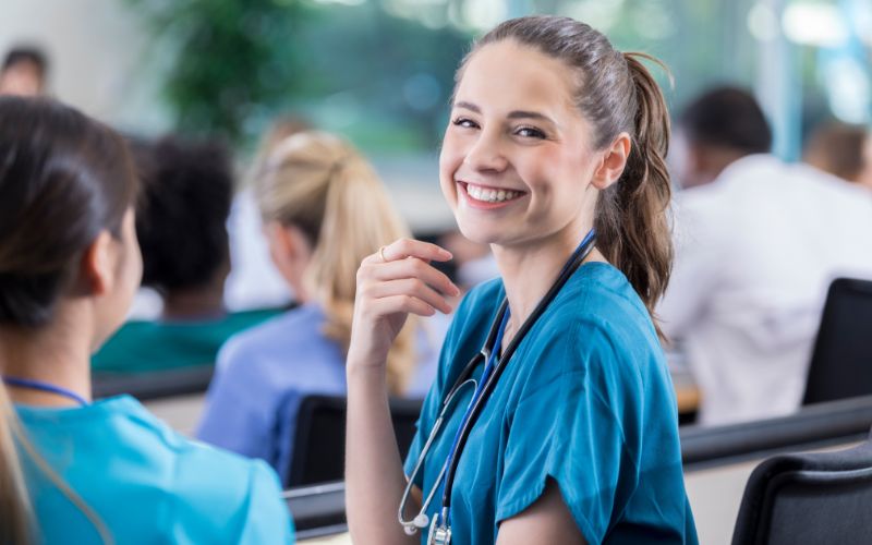 Na zdjęciu znajduje się uśmiechnięta studentka medycyny ubrana w niebieski fartuch i zawieszony na szyi stetoskop. Znajduje się w sali na uniwersytecie.