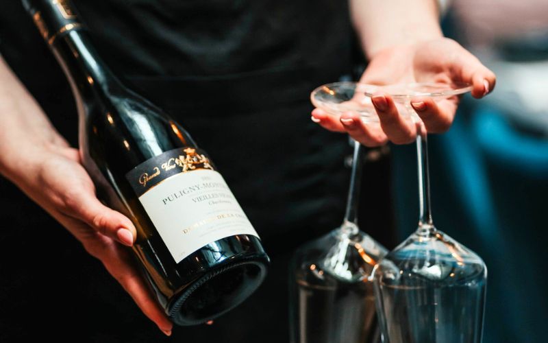 Zdjęcie przedstawia osobę, która w jednej ręce trzyma butelkę wina, natomiast w drugiej dłoni 2 kieliszki wina