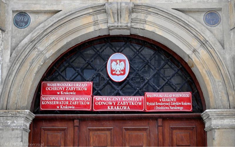 Zdjęcie przedstawia drzwi wejściowe do budynku Urzędu Ochrony Zabytków w Krakowie. Przy drzwiach znajdują się tabliczki informacyjne.