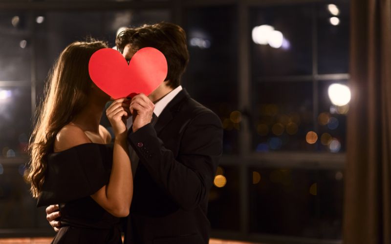 Zdjęcie przedstawia parę osób, która zasłania swoją twarz kartonem w kształcie serca